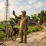 Postino rurale indiano misura il segnale delle reti mobili con smartphone in un villaggio, torre telecom sullo sfondo - accordo TRAI Department of Posts