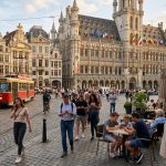 Piazza storica europea affollata al tramonto con persone che usano lo smartphone e bandiere di diversi Paesi UE: il rapporto BEREC sul consolidamento delle telco europee