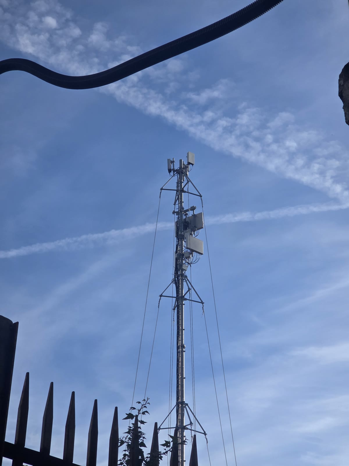 Il sito carrato Vodafone acceso per Lucca Comics 2026: palo telescopico con tiranti, antenne e unitร radio in cima, visto da un cancello, cielo azzurro